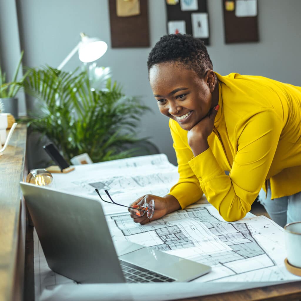 Young woman leaning over desk with blueprint looking at laptop